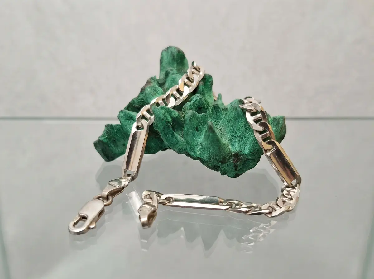 Silbernes Gliederarmband mit Plättchen, das dekorativ über einen rauen grünen Malachit-Stein auf einer Glasfläche drapiert ist