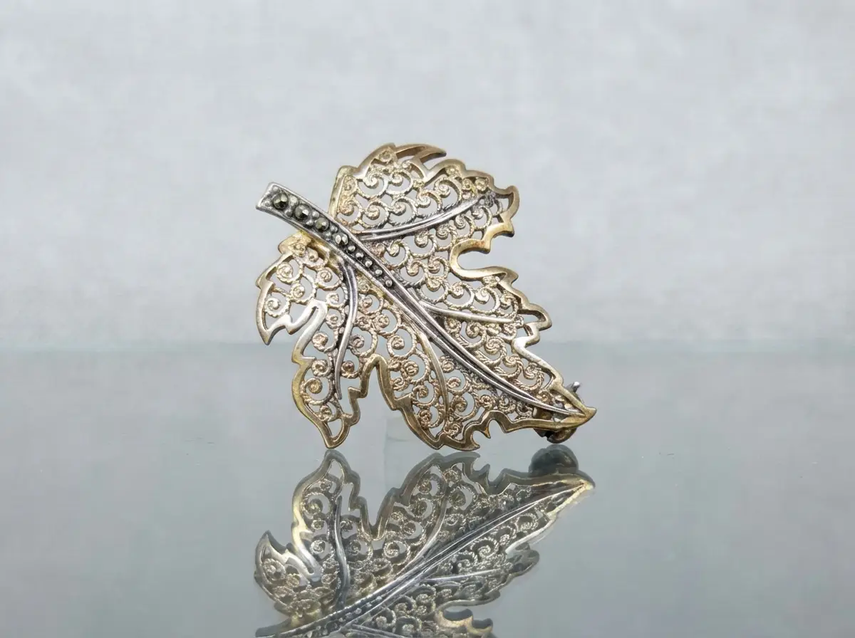 Zweifarbige Brosche in Blattform mit filigranen Verzierungen und Markasit-Steinen auf dem Stiel, stehend auf einer spiegelnden Oberfläche.