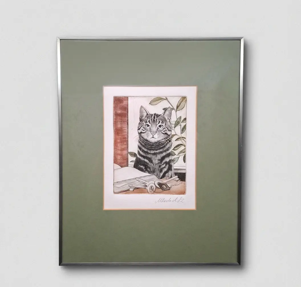 Gerahmte Radierung einer getigerten Katze, die hinter einem Schlüsselbund und einem Buch sitzt, präsentiert in einem olivgrünen Passepartout und silbernem Rahmen an einer weißen Wand.