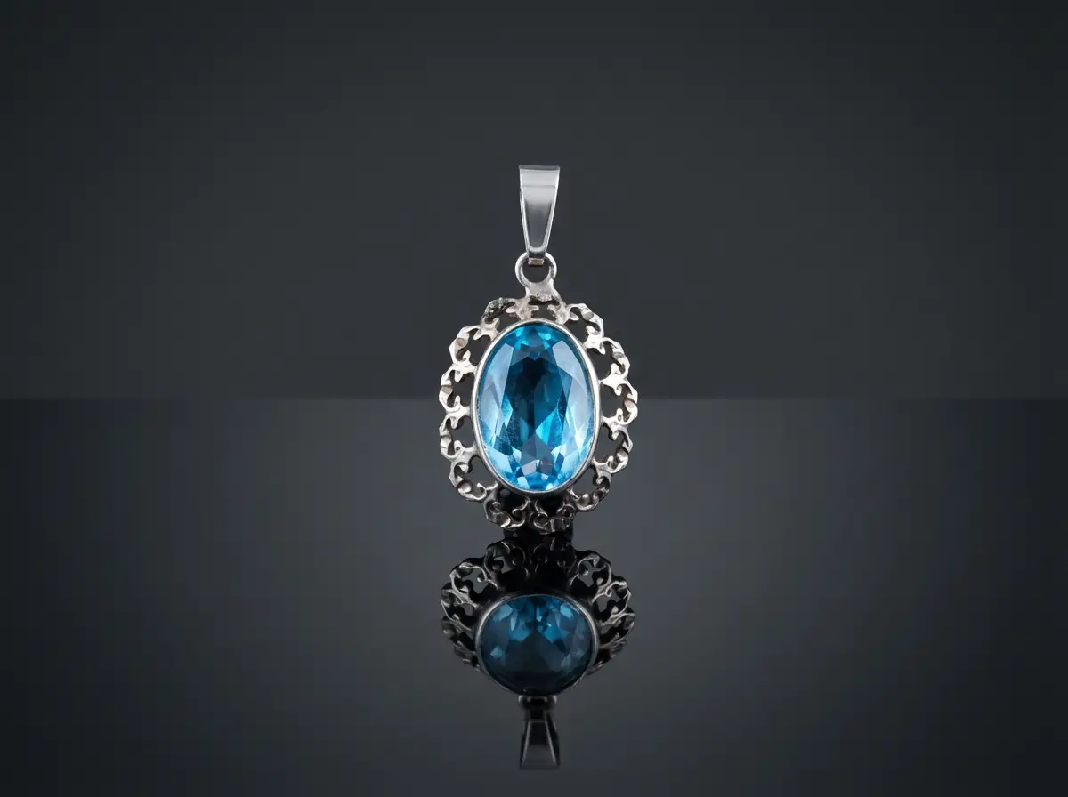 Filigraner Silberanhänger mit einem facettierten, ovalen blauen Edelstein auf schwarzem, spiegelndem Untergrund.