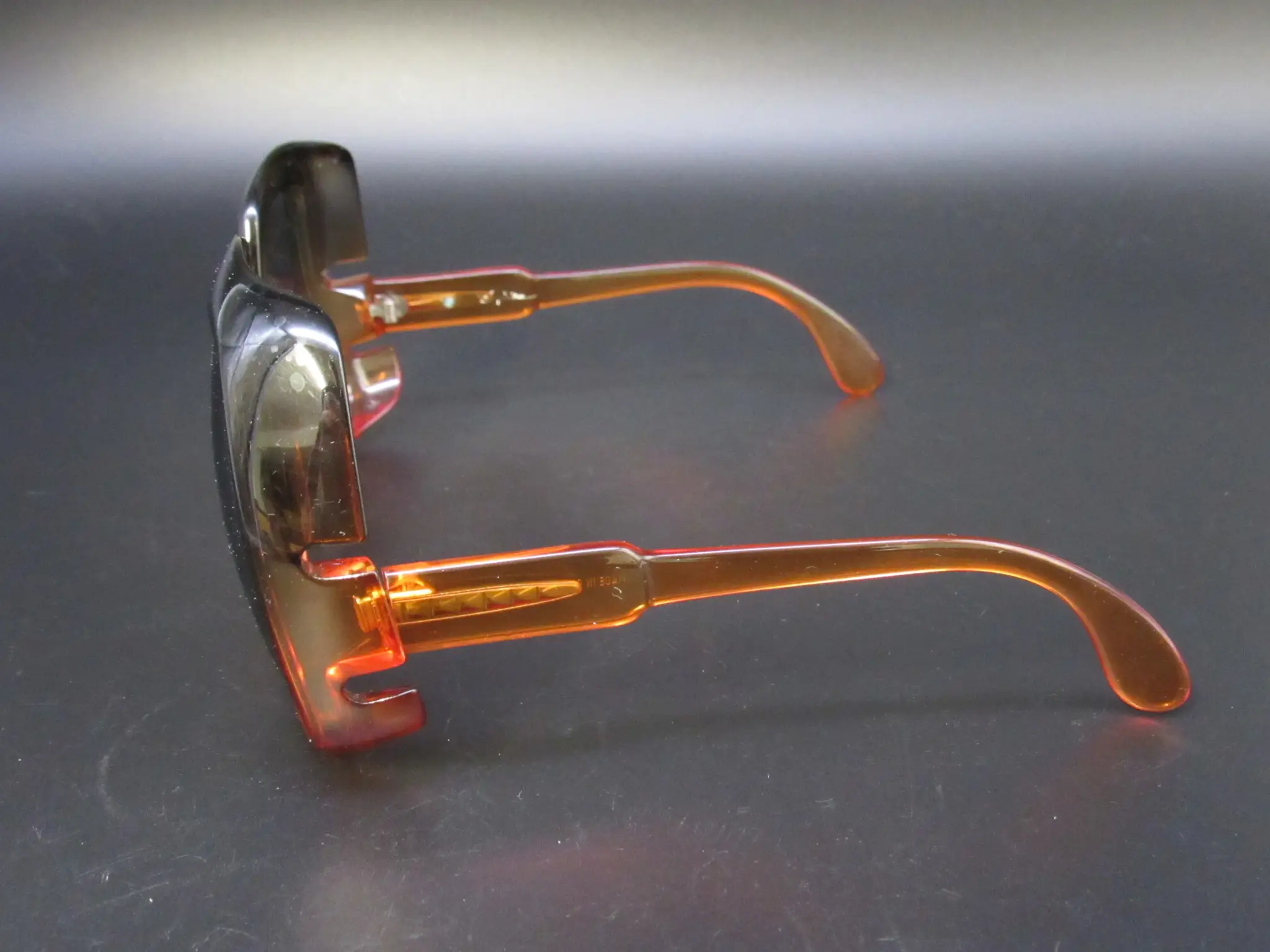 Seitenansicht einer Vintage-Sonnenbrille mit dunklen Gläsern und breiten, transparenten orangen Bügeln vor dunklem Hintergrund