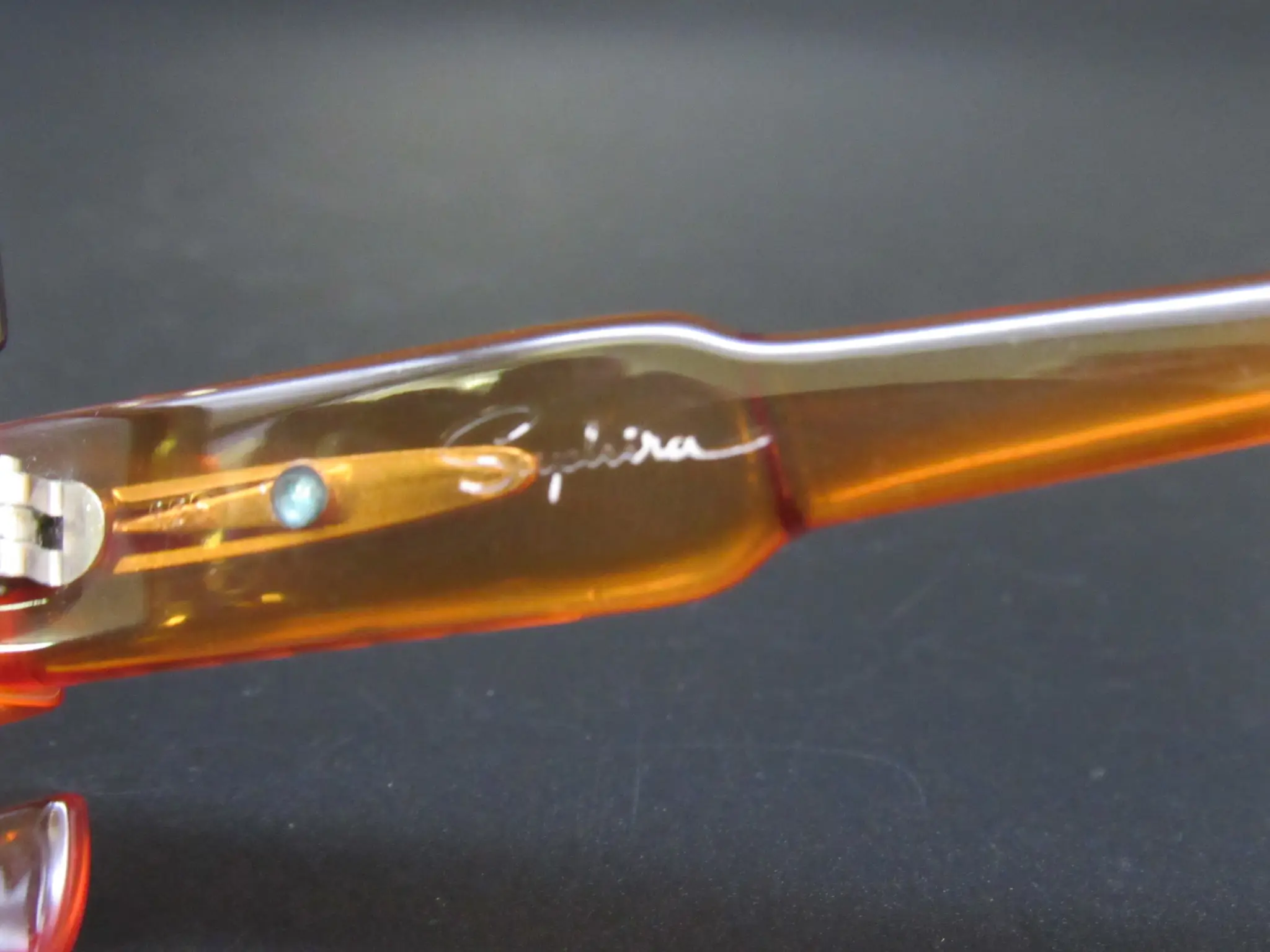 Nahaufnahme eines bernsteinfarbenen Saphira Brillenbügels mit weißem Schriftzug und sichtbarem Scharnier auf dunklem Hintergrund
