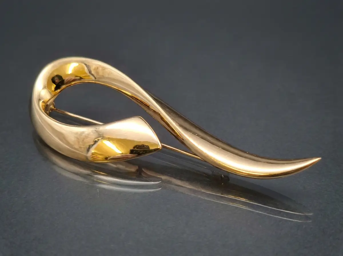 Glänzende goldene Brosche in geschwungener, abstrakter Form auf dunklem Untergrund mit Spiegelung