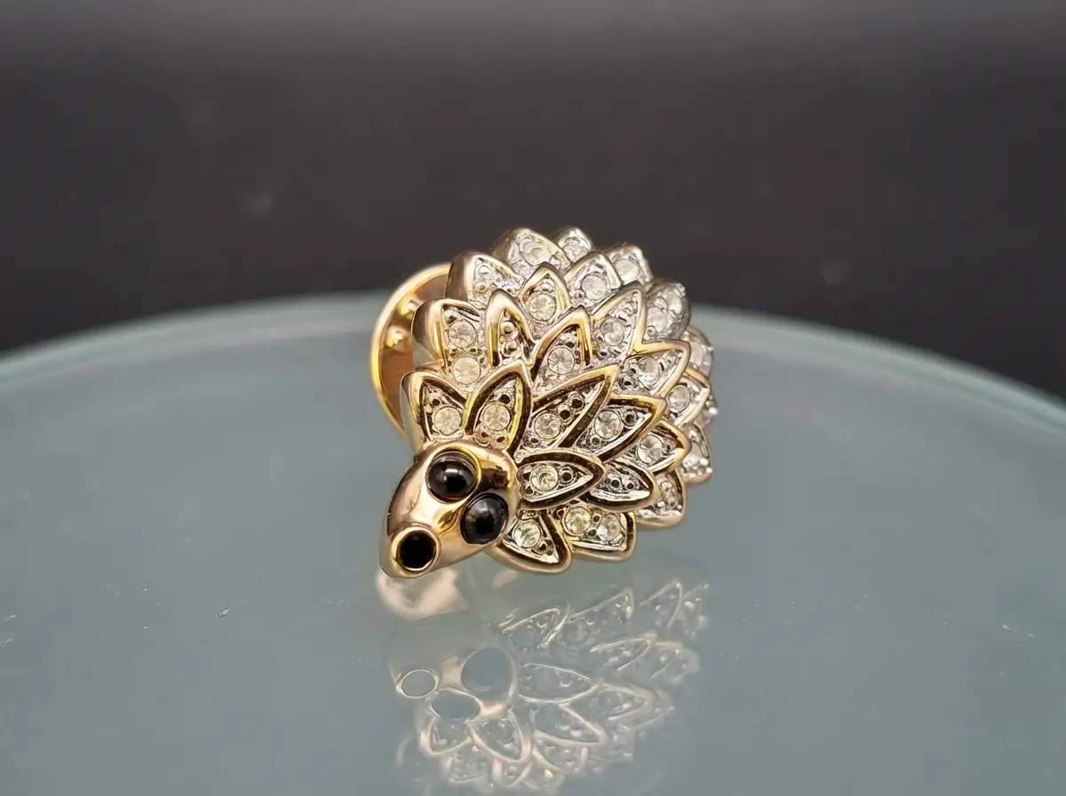 Goldene Anstecknadel in Igel-Form, besetzt mit klaren Strasssteinen und schwarzen Augen auf einer spiegelnden Oberfläche