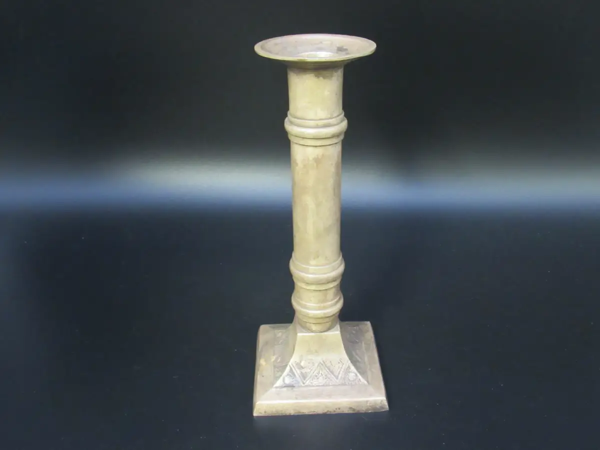 Messing-Kerzenständer mit säulenförmigem Schaft und quadratischem Fuß mit geometrischer Gravur vor schwarzem Hintergrund