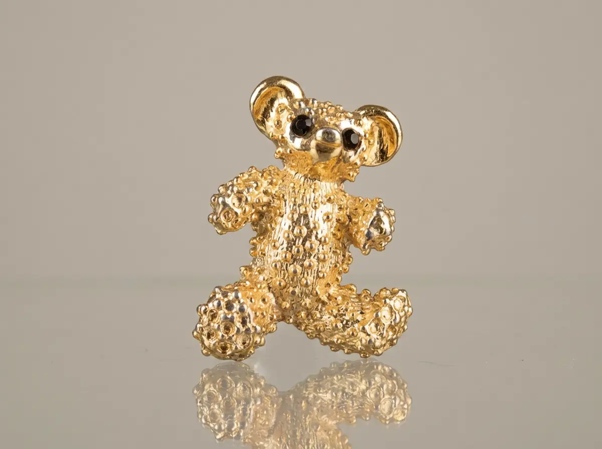 Goldene Brosche in Form eines Teddybären mit texturierter, genoppter Oberfläche und dunklen Augensteinen vor neutralem Hintergrund mit Spiegelung