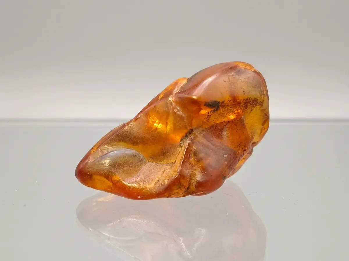 Nahaufnahme eines orange-braunen Bernsteins mit sichtbaren internen Strukturen und Rissen, der auf einer reflektierenden Oberfläche vor neutralem Hintergrund liegt