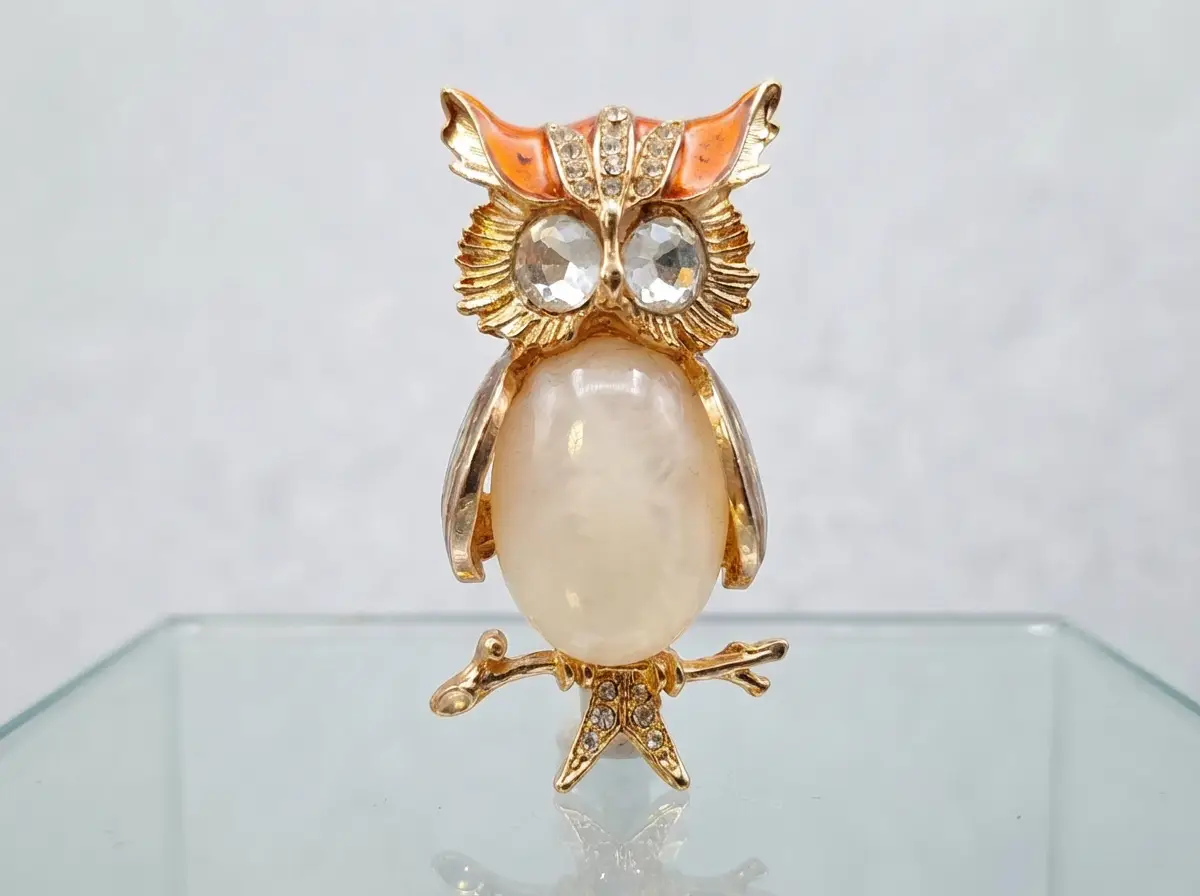 Goldfarbene Brosche in Eulenform mit großen Strass-Augen, orangefarbenen Akzenten am Kopf und einem großen, ovalen, cremefarbenen Stein als Körper