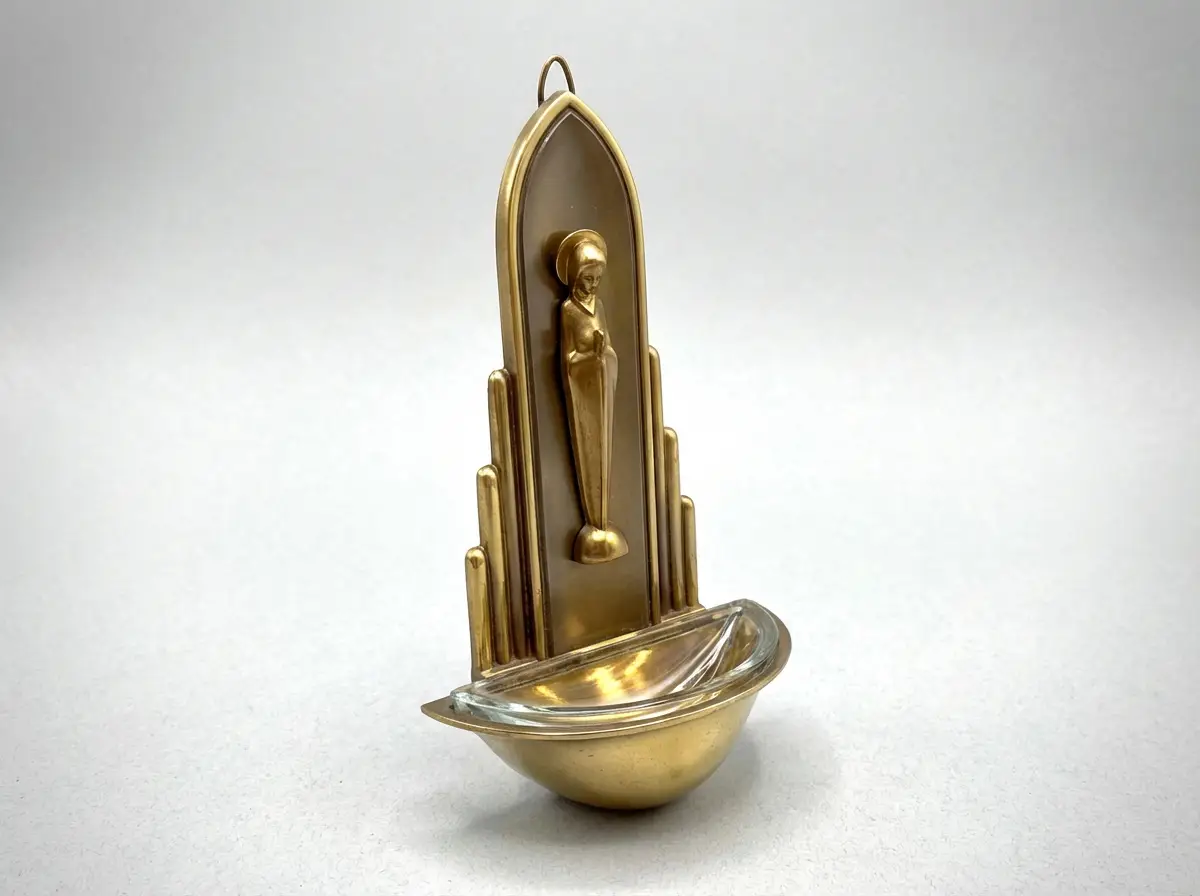 Messing-Weihwasserkessel im Art-Deco-Stil mit stilisierter Madonna-Relieffigur, gestuften Seiten und Glaseinsatz vor weißem Hintergrund