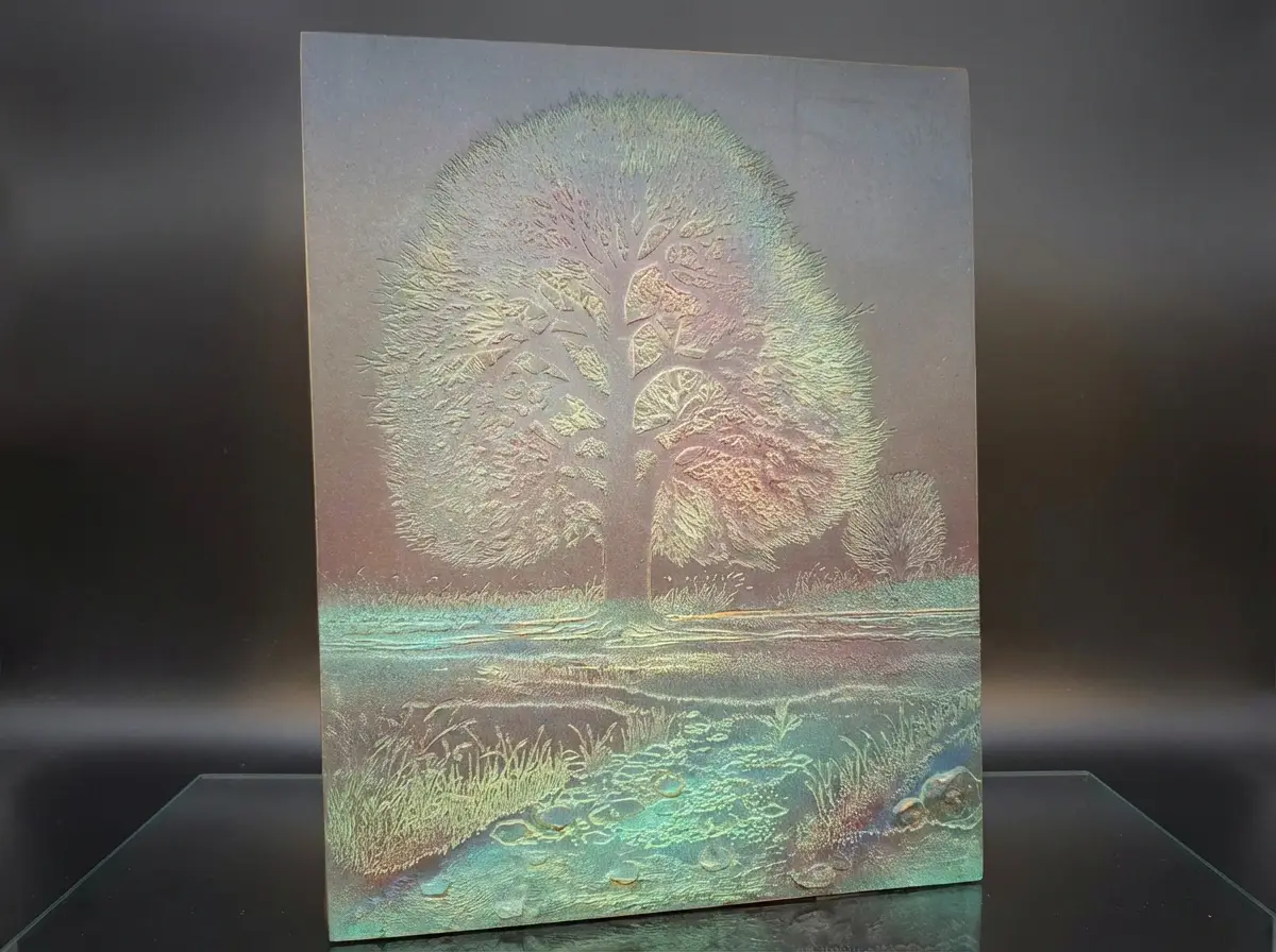 Rechteckiges Metallrelief mit einer detaillierten Gravur eines Baumes in einer Landschaft, schimmernd in irisierenden Grün- und Kupfer-Tönen vor schwarzem Hintergrund