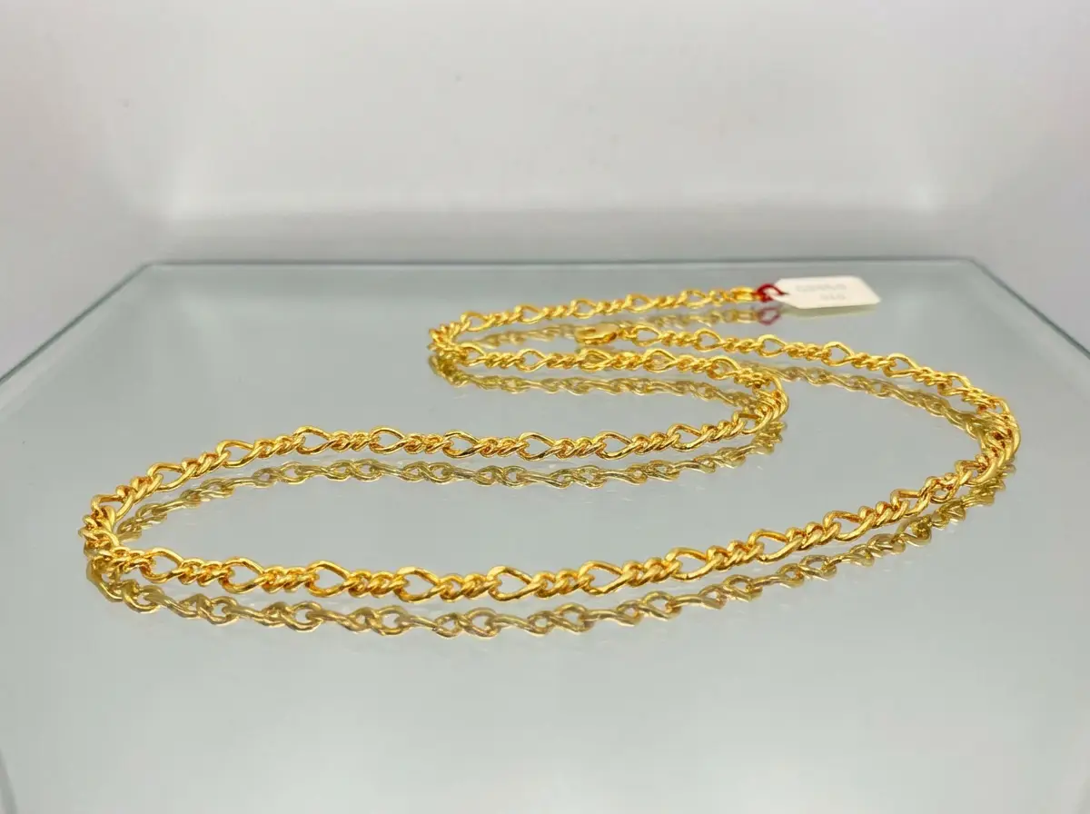 Eine goldene Gliederkette, die geschwungen auf einer spiegelnden Glasoberfläche liegt, mit einem sichtbaren weißen Etikett am Verschluss im Hintergrund.