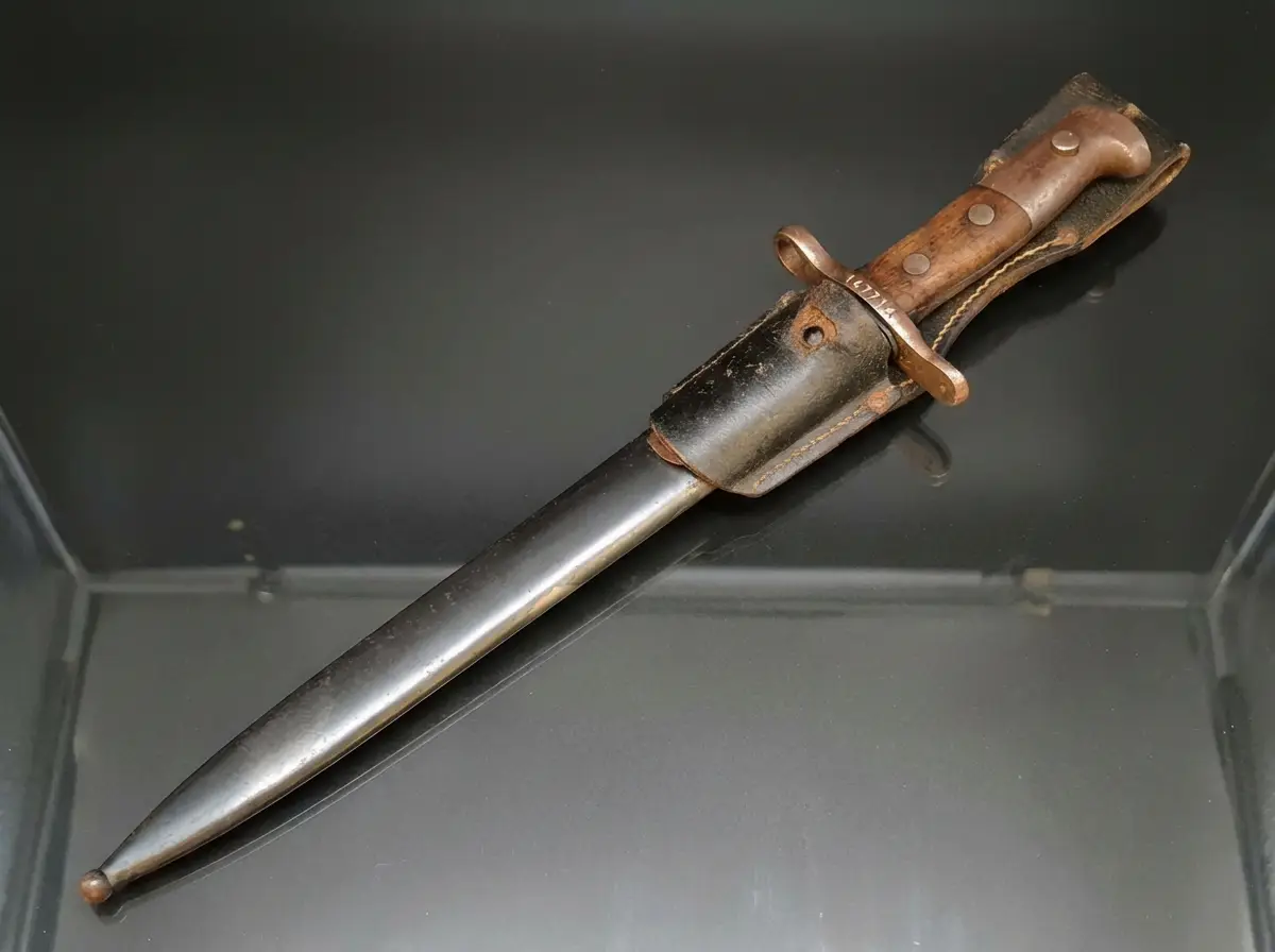 Antikes militärisches Bajonett mit Holzgriffschalen in schwarzer Metallscheide samt Lederkoppelschuh auf dunklem Untergrund