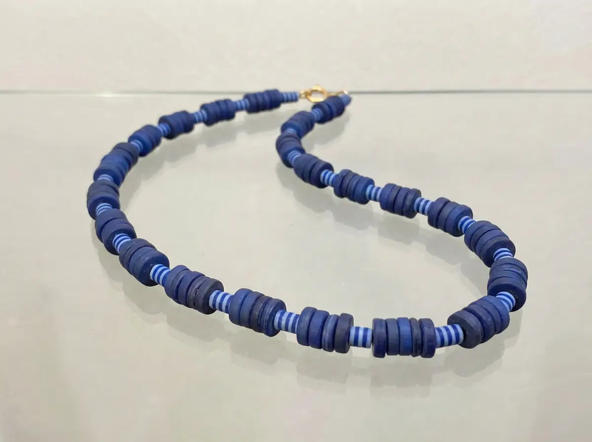 Dunkelblaue Halskette aus flachen Scheibenperlen mit hellblauen gestreiften Zwischenperlen und goldenem Verschluss auf hellem Untergrund
