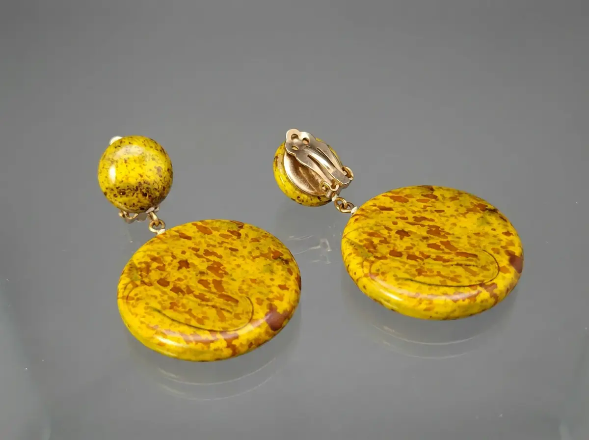 Paar runde gelbe Clip-Ohrringe mit brauner Musterung und goldfarbenem Verschluss auf grauem Untergrund