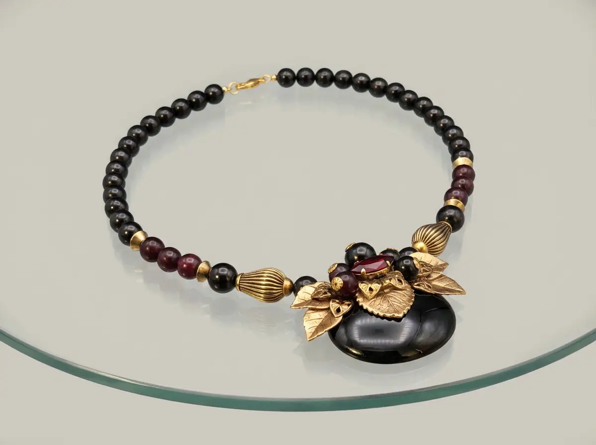 Elegante Halskette aus schwarzen Perlen mit goldenen Blatt-Elementen, dunkelroten Akzenten und einem großen schwarzen Anhänger auf einer Glasfläche