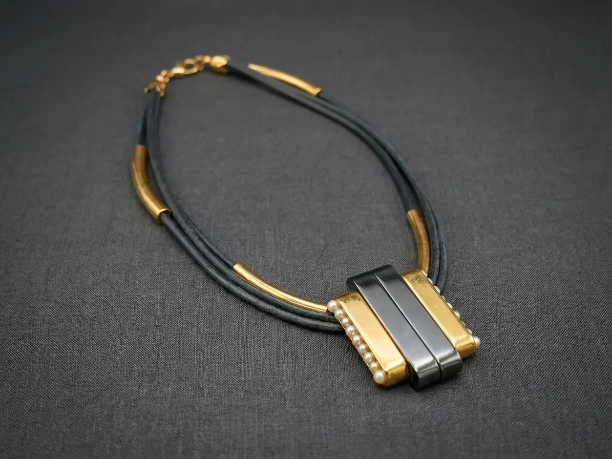 Mehrreihige Halskette in Schwarz und Gold mit rechteckigem Anhänger, der mit kleinen Perlen besetzt ist, auf dunklem Hintergrund