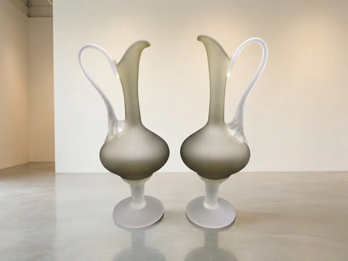 Zwei symmetrisch aufgestellte bauchige Glasvasen mit schlankem Hals und geschwungenem Henkel aus mattem Glas auf einem hellen spiegelnden Boden