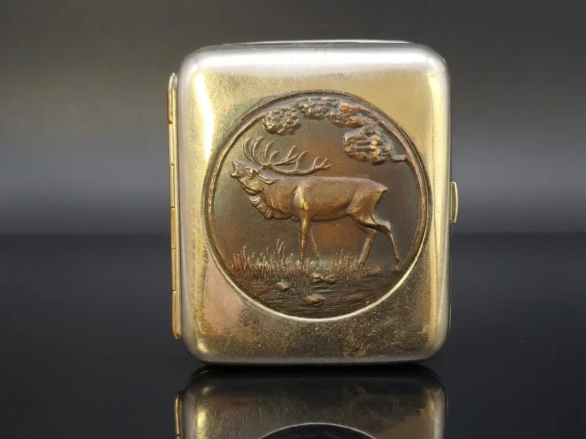 Ein kleines, rechteckiges Metalletui, wahrscheinlich eine Pillendose oder ein Schnupftabakdöschen, mit einer kreisförmigen Prägung eines stehenden Hirsches in einer Landschaft.