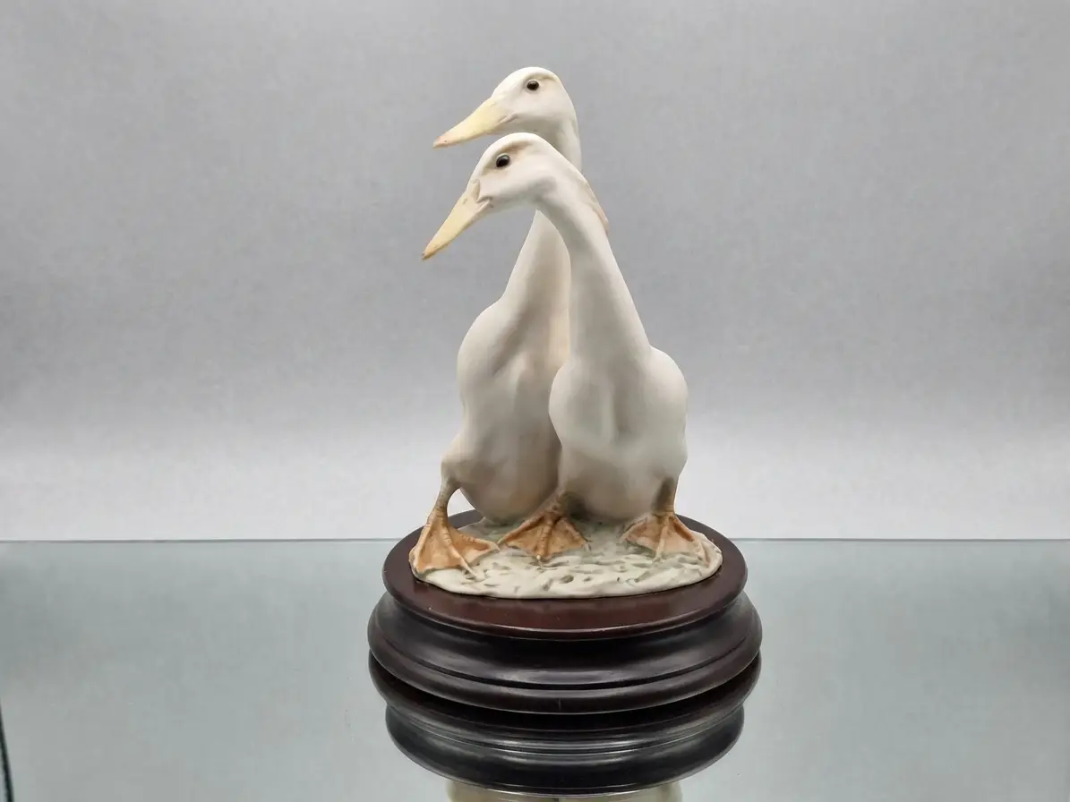 Eine dekorative Porzellanfigur, die zwei weiße Enten darstellt, welche auf einem dunklen runden Holzsockel stehen.
