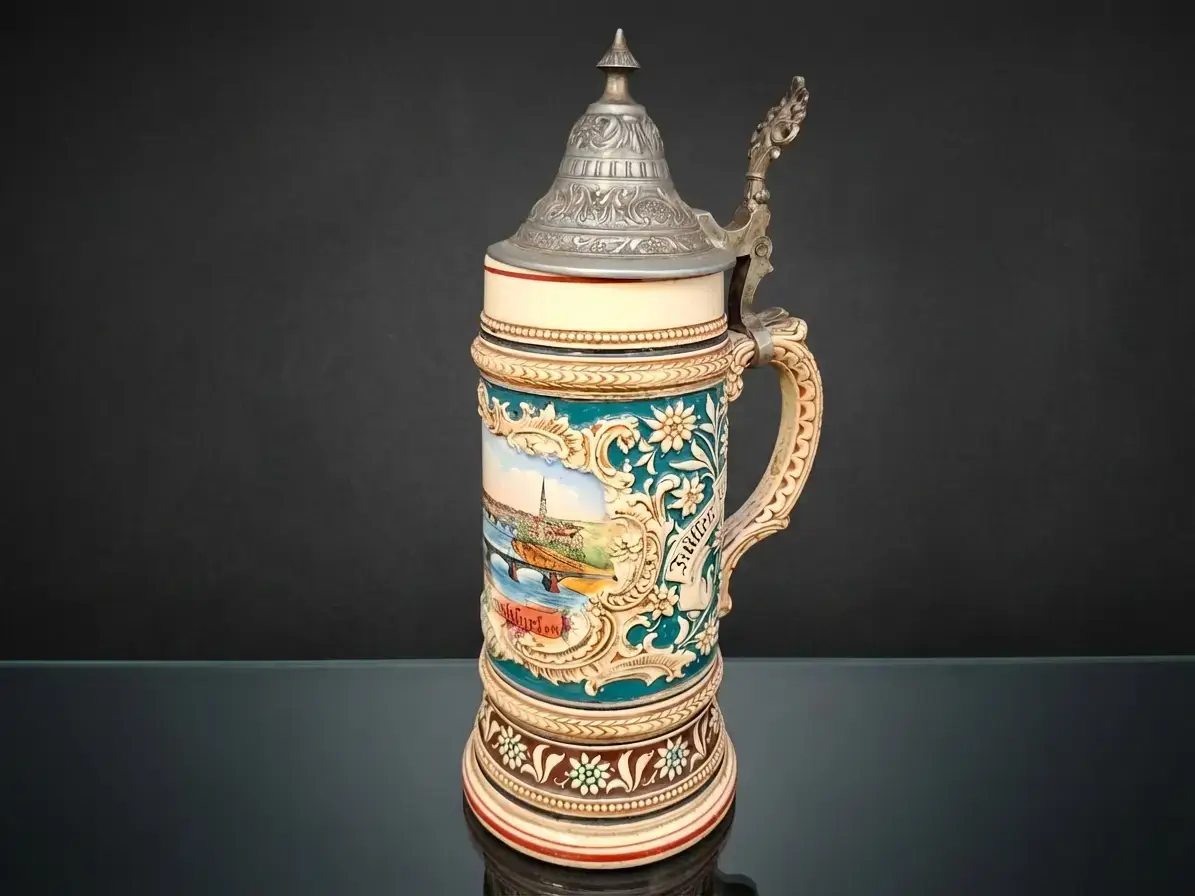 Reich verzierter traditioneller Keramik-Bierkrug mit Zinndeckel, floralen Mustern und einem Stadtmotiv vor dunklem Hintergrund.