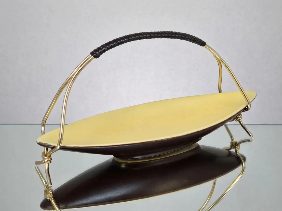 Längliche Vintage-Schale im Mid-Century-Design, innen gelb, außen braun, mit einem geschwungenen Messingbügel. Das Objekt spiegelt sich auf einer glatten Oberfläche.