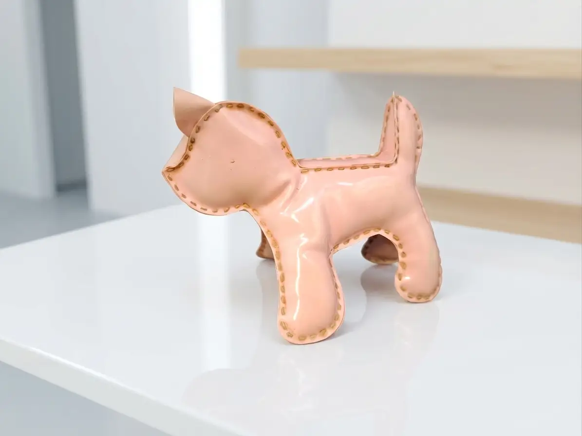 Eine kleine, glänzende rosa Dekofigur in Form eines Hundes mit braunen Ziernähten, die auf einer weißen Hochglanzfläche steht.